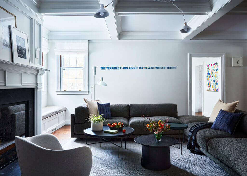 A traditional family room with a classic fireplace, coffered ceiling, and modern furniture, including a black sectional, a trio of tables, and curvy swivel chairs from Design Within Reach.