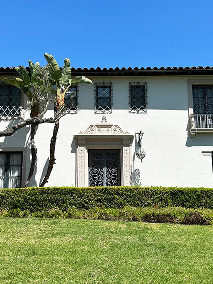 Historic architecture in Los Angeles shown in a stucco home with a clay tile roof, palm tree, and lush green landscaping.